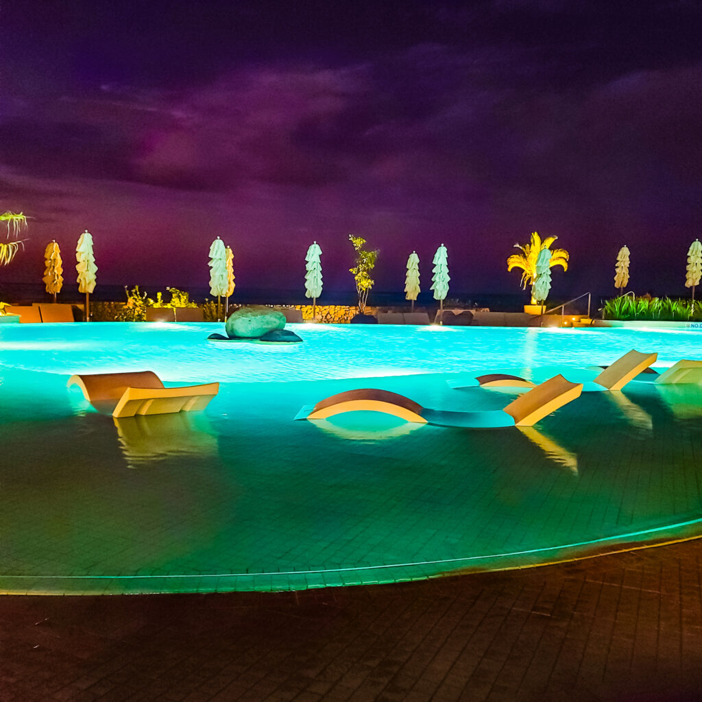 Solid, white pool loungers halfway sticking out above the surface of an illuminated pool against a very dark purple night sky