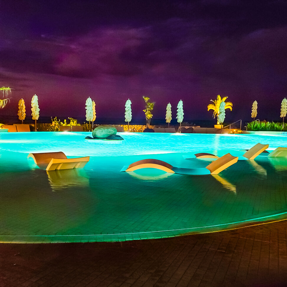 Solid, white pool loungers halfway sticking out above the surface of an illuminated pool against a very dark purple night sky