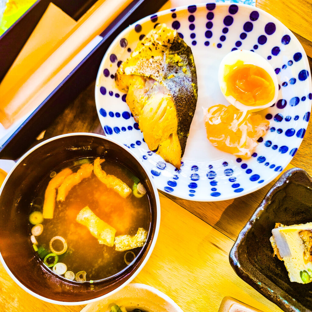 An above view of a fat fish filet marinated in saikyo-yaki miso with a hard-boiled egg on a white dish with vertical columns of blue dots around the brim. A black bowl of miso soup on the left side.