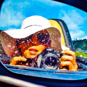 Passenger-side mirror with a reflection of a woman with floppy hat and shades in a convertible holding a camera with a Hawaiian hillside in the background.