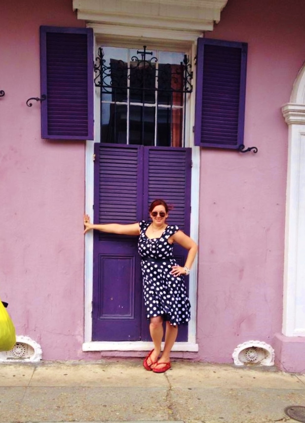 Me in a knee-length black and white polka dot dress with red thong sandals standing in front of purple shutters closed over a tall window with wrought iron on a light purple house in the French Quarter.