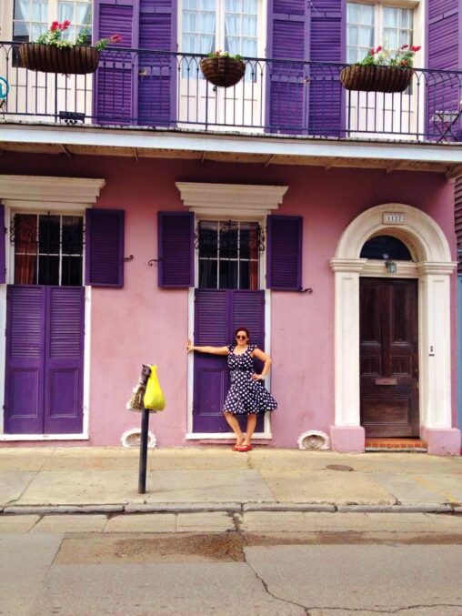 Me in a knee-length black and white polka dot dress with red thong sandals standing in front of purple shutters closed over a tall window with wrought iron on a light purple house in the French Quarter.
