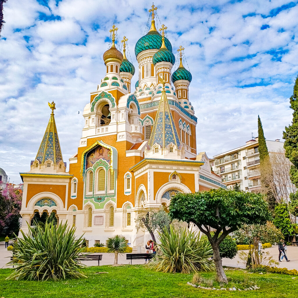Russian Orthodox Cathedral of Nice . Orange brickwork with cream-colored brick trim and lavish aqua green embellishments and inlays, topped with a large aqua green pointed dome with 3 smaller ones in front of it, and an orange one on the left. Two blue steeples with gold embellishments in the front and left side.