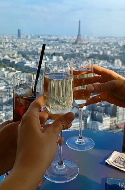 3 hands holding 3 champagne flutes, one with Coke, joined together in front of a daytime city view of Paris with the Eiffel Tower in the distance.